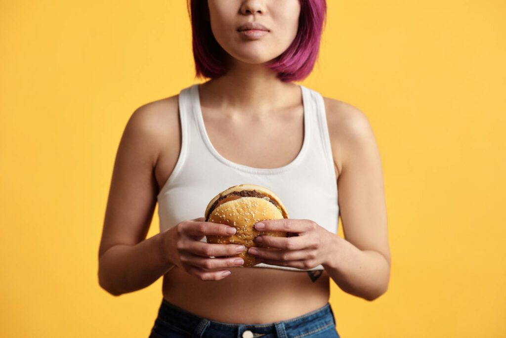 Young woman with pink hair holding a burger, set against a yellow backdrop.